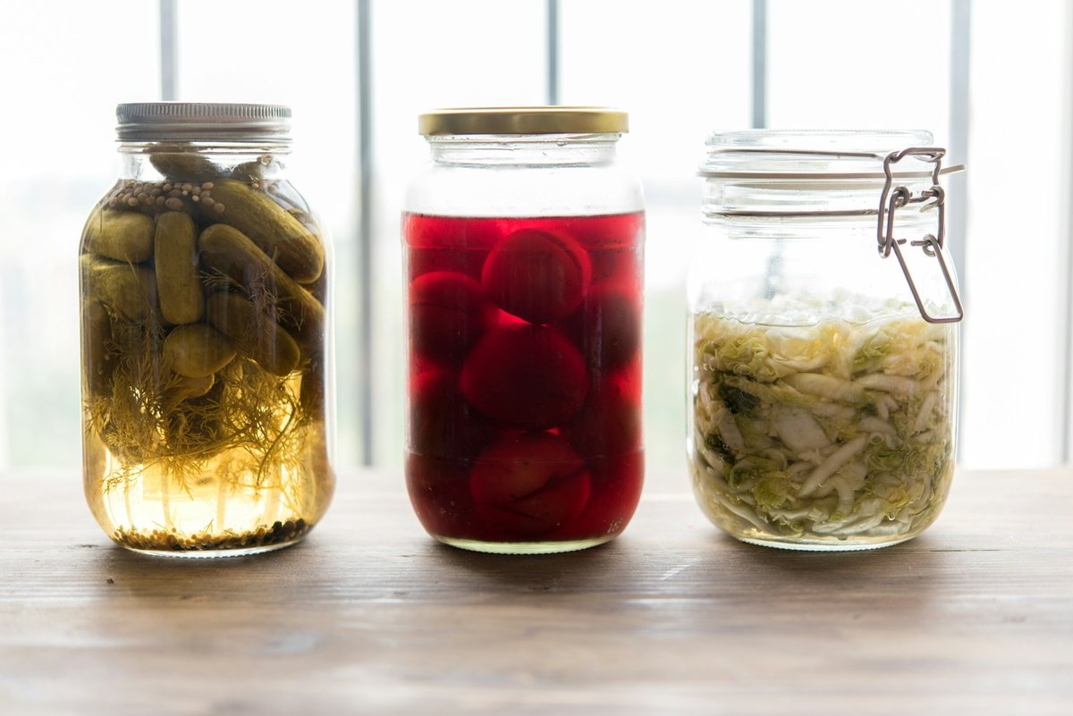Trois bocaux de cornichons, de betteraves et de chou lactofermentés devant une fenêtre